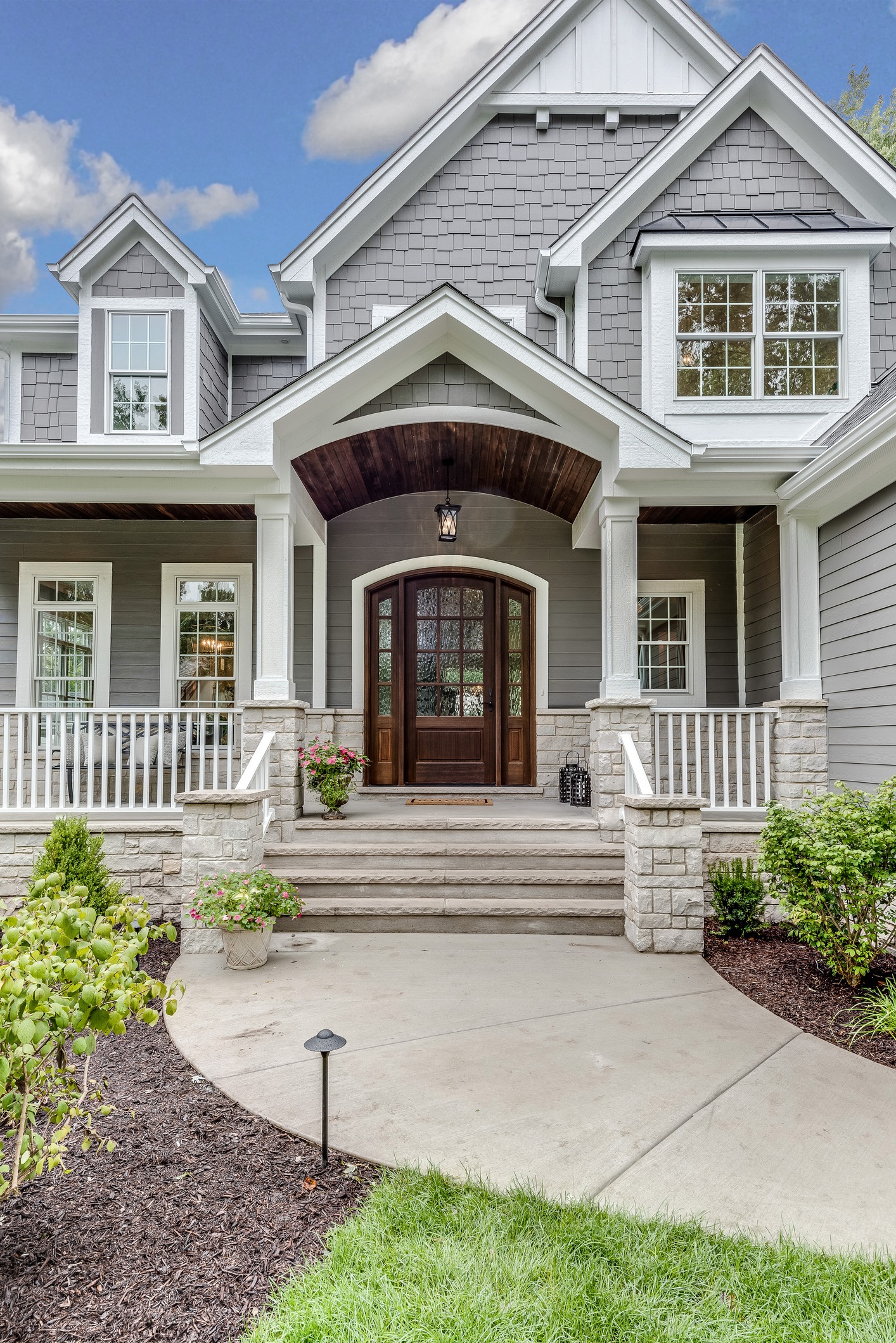 Front view of a modern gray house with white trim, a covered porch, large wooden double doors, stone pillars, and landscaped greenery along a curved walkway under a partly cloudy sky.