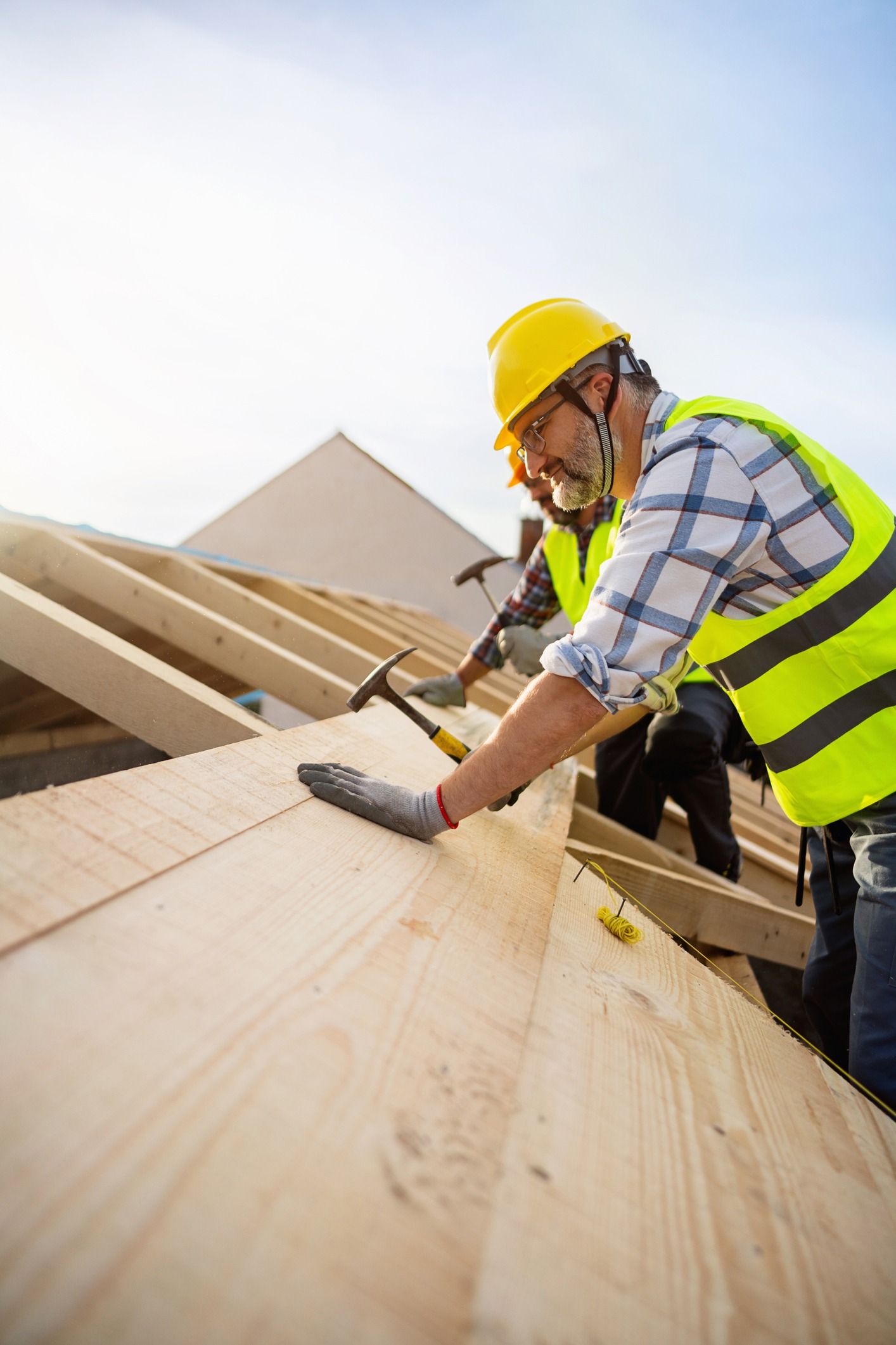 A construction worker in a yellow safety vest and helmet uses a hammer to secure wooden beams on a roof under construction, with blue sky in the background.