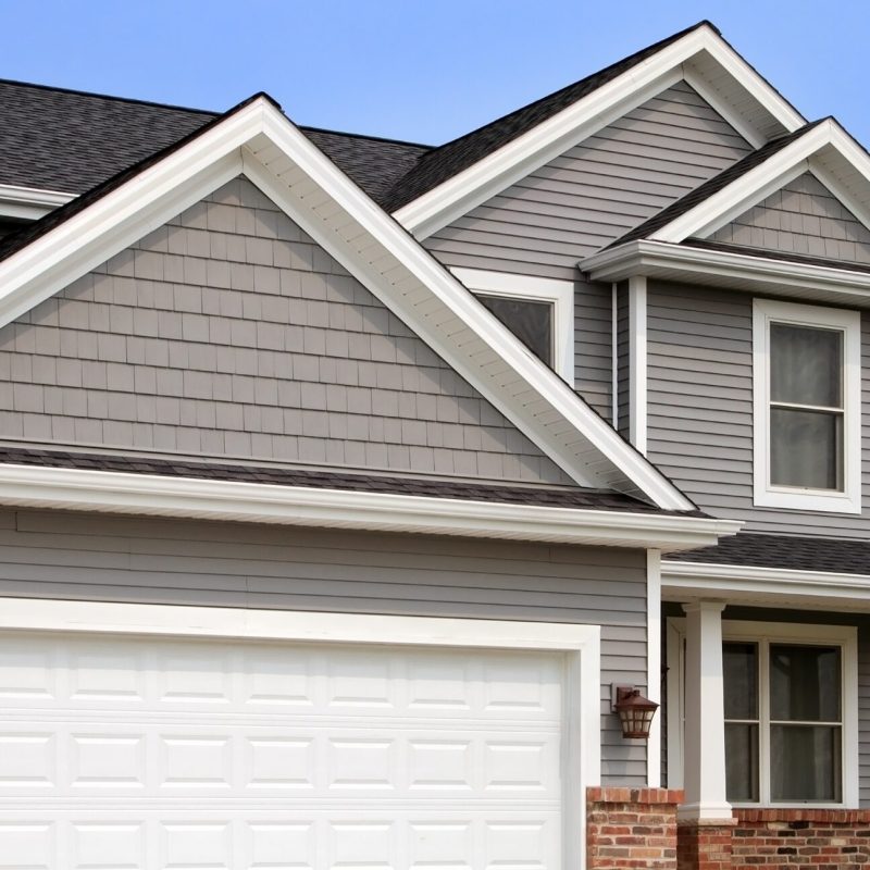 Modern suburban house with gray siding, white trim, large windows, and a white double garage door. The roof is dark gray and gabled, with brick accents at the base of the exterior walls.