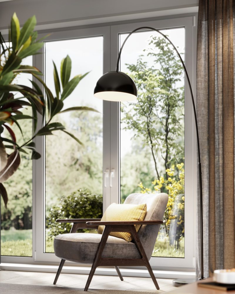 A cozy reading nook with a modern armchair, yellow cushion, and arc floor lamp sits by large glass doors overlooking a green garden with trees and bushes. Sunlight filters through, creating a warm, inviting atmosphere.