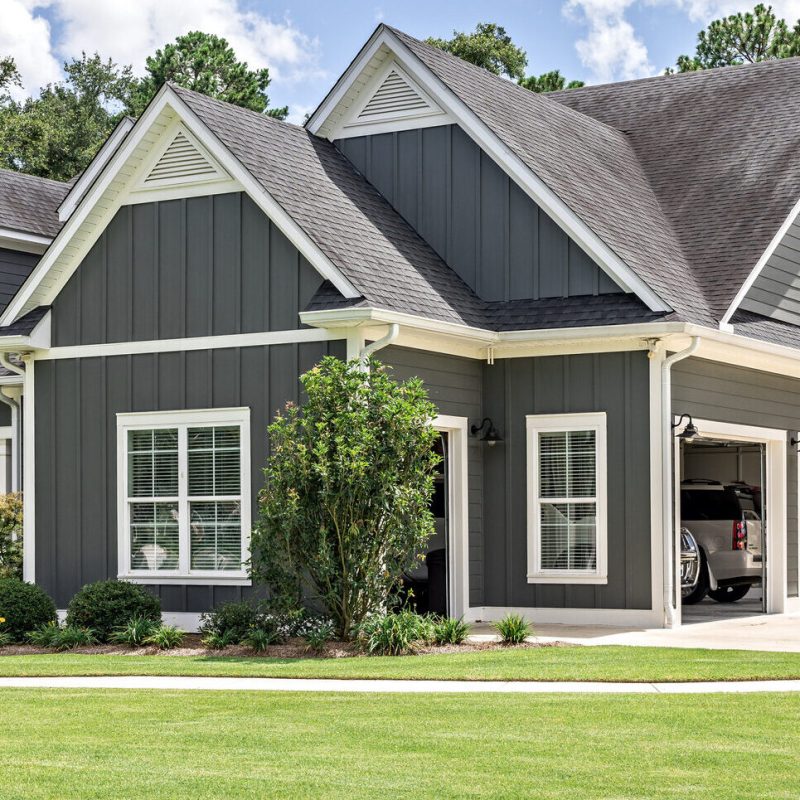 A modern, two-story house with gray siding, white trim, and a large attached garage. The home is surrounded by a green lawn, bushes, and trees, with a driveway leading to the garage. An American flag hangs by the garage.
