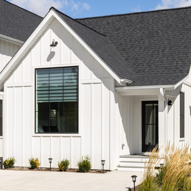 A modern white house with board and batten siding, large black-framed window, black roof, and small front porch. Minimal landscaping with shrubs and ornamental grasses lines a concrete path.