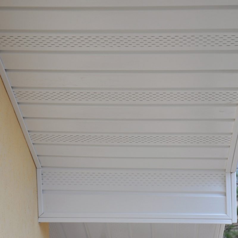 Close-up view of a house's soffit and fascia with white vented panels under the roof overhang, attached to a yellow exterior wall. Pine tree branches are visible on the right side.