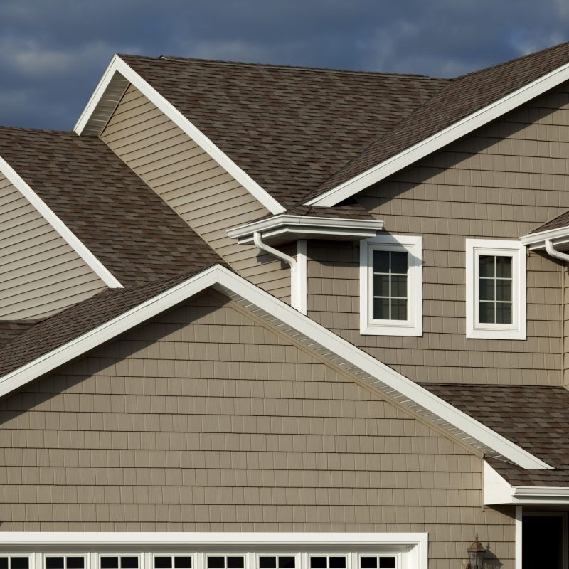 Close-up view of a modern house exterior with taupe-colored siding, white trim, and multiple intersecting gable roofs, set against a cloudy sky.