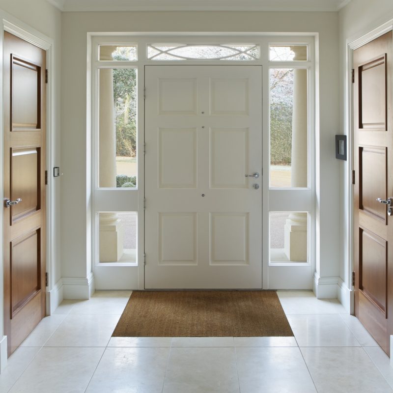 A bright, tidy entryway with a large white front door, sidelights, two wooden doors on either side, tiled floor, and a brown doormat. Sunlight streams through the glass panes, illuminating the space.