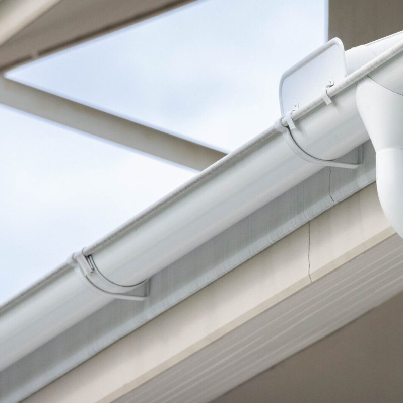 Close-up of clean, white rain gutters and downspouts attached to the edge of a house roof against a light blue sky. The gutter system is neatly installed with brackets and connectors visible.