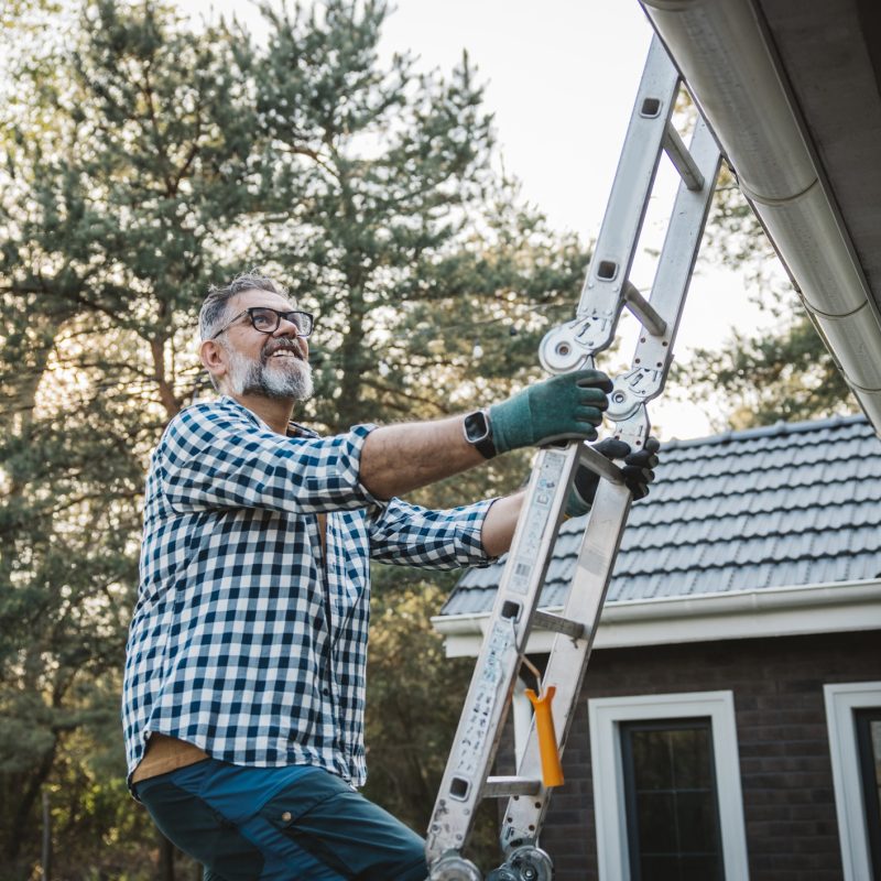 A man wearing glasses and gloves stands on a ladder, cleaning or inspecting the gutter of a house with a tiled roof, surrounded by trees, on a sunny day.