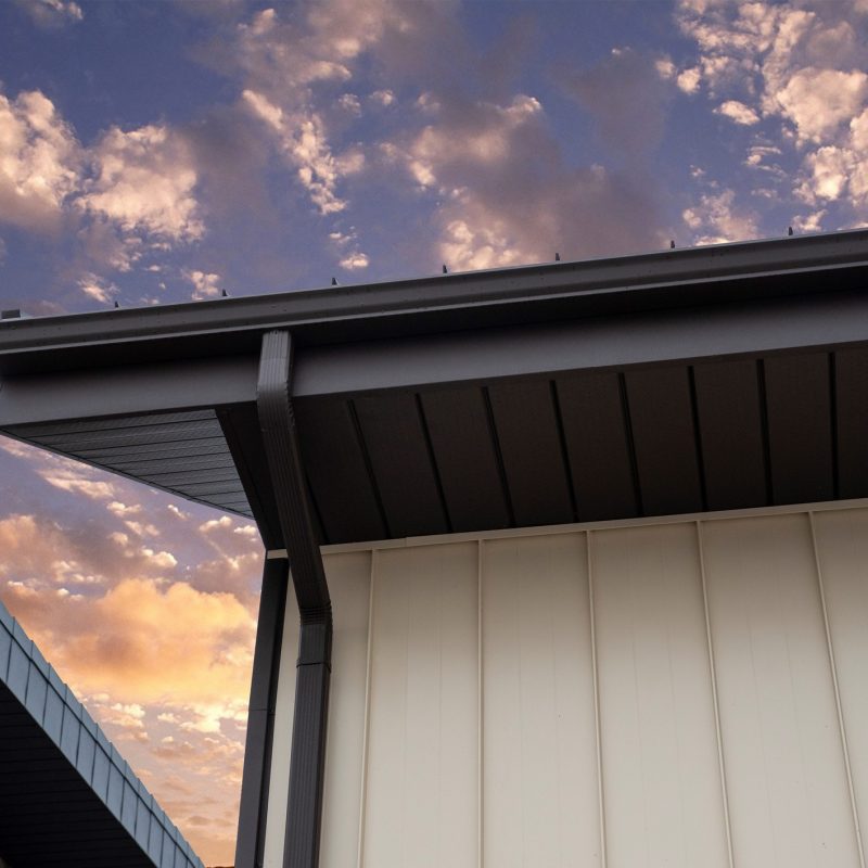 Close-up view of modern building roofs with metal gutters, set against a dramatic sky with scattered clouds at sunset or sunrise. The architecture features clean lines and a mix of cream and blue-gray panels.