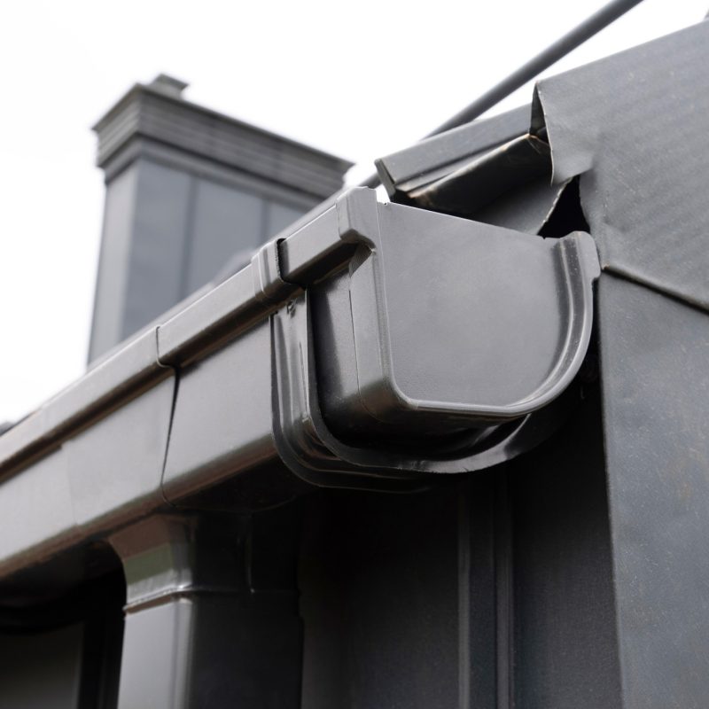 Close-up of a modern black rain gutter system attached to the edge of a roof on a building with dark siding, with a downspout visible and a chimney in the background.