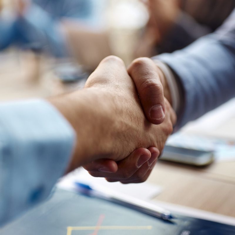 Two people in business attire shaking hands across a table, suggesting a successful agreement or partnership, with blurred office materials and colleagues in the background.